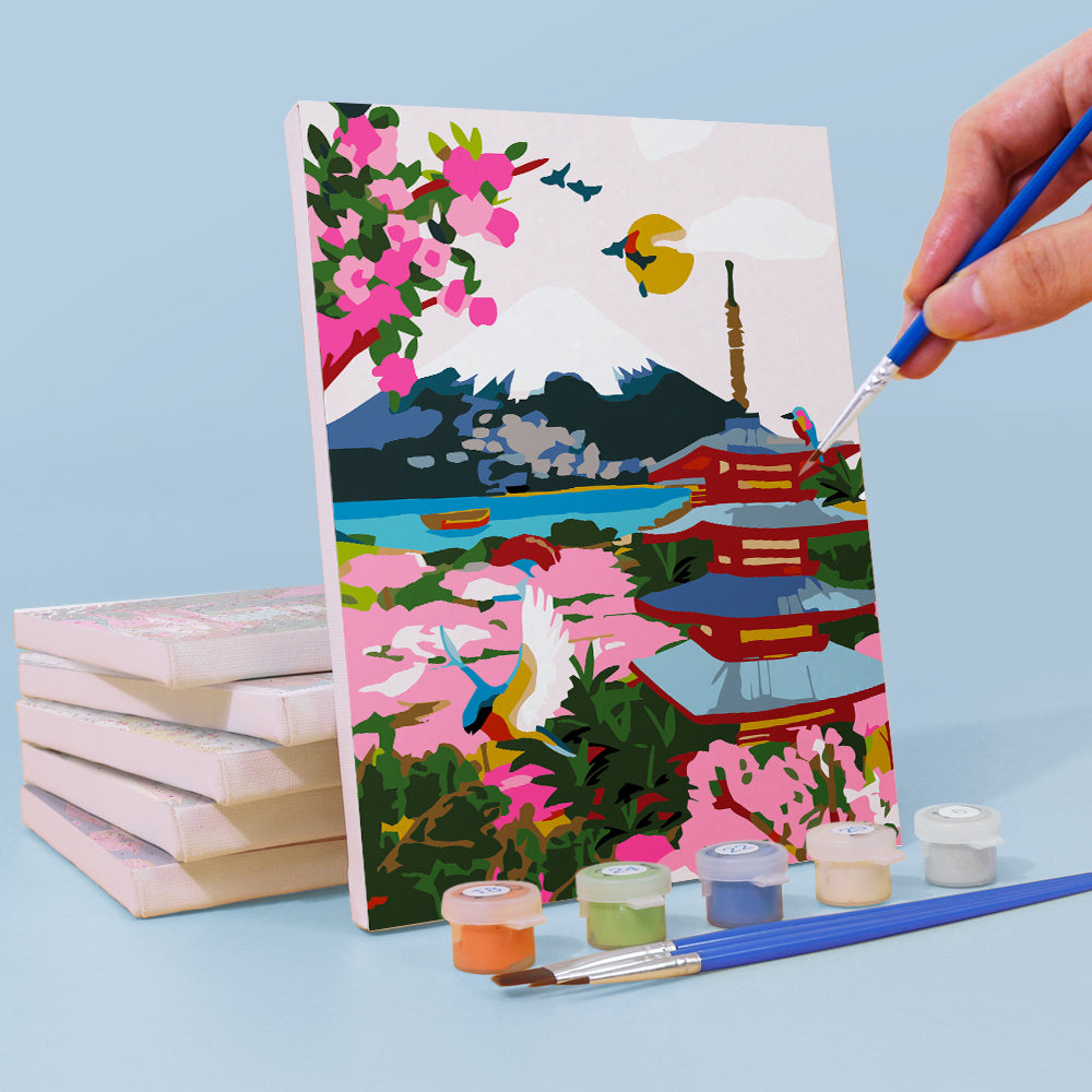 Painting of a scenic landscape with cherry blossoms and a pagoda, with a hand holding a paintbrush. Mini Painted Journeys 6 Pack Paint by Numbers