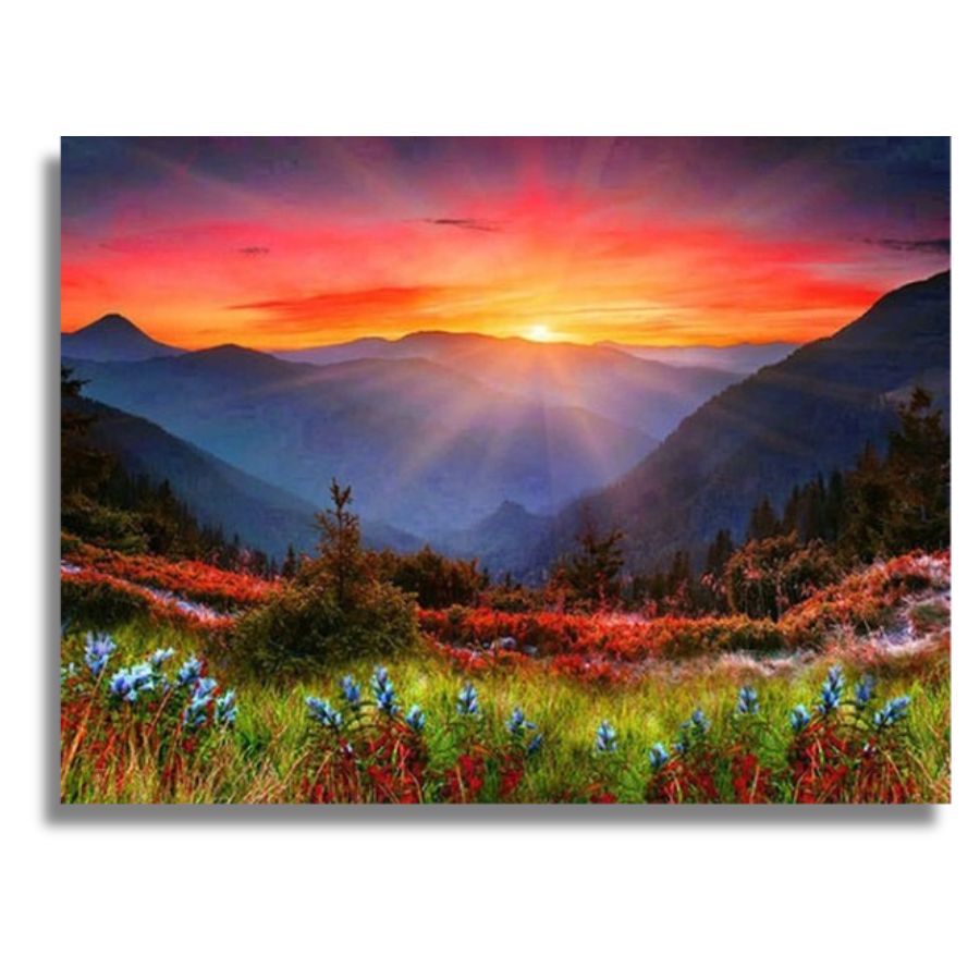 A rectangular wall art print or canvas showing a mountain landscape at sunset, with blue mountains in the distance, a glowing orange and pink sky, and wildflowers in green, red, and blue in the foreground. The print is photographed against a light gray background with a soft drop shadow. Burning Sunset in the Mountains Canvas - Paint by numbers