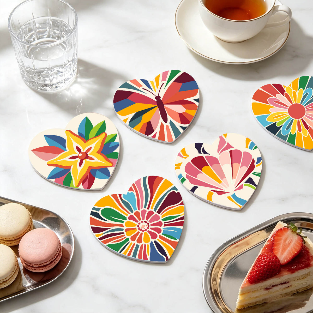 Six heart-shaped coasters with colorful, vibrant abstract and floral patterns arranged on a white marble tabletop alongside a glass of water, a cup of tea, macarons, and a strawberry cake slice. Colorful Kaleidoscope Coasters - 6 Pack Paint by Numbers Lifestyle
