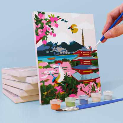 Painting of a scenic landscape with cherry blossoms and a pagoda, with a hand holding a paintbrush. Mini Painted Journeys 6 Pack Paint by Numbers