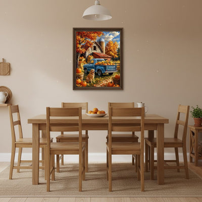 A framed autumn landscape painting hangs on a beige wall above a wooden dining table and chairs. The painting depicts a blue pickup truck parked in a fall yard with pumpkins, orange trees, and a barn. Autumn Harvest Truck - Paint by numbers lifestyle