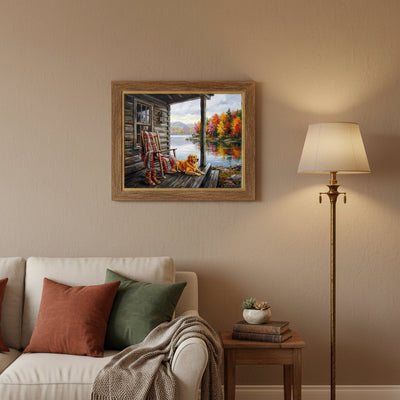 A framed landscape painting of a cozy cabin porch with a dog by a lake, displayed on a beige wall above a white sofa in a styled living room setting.