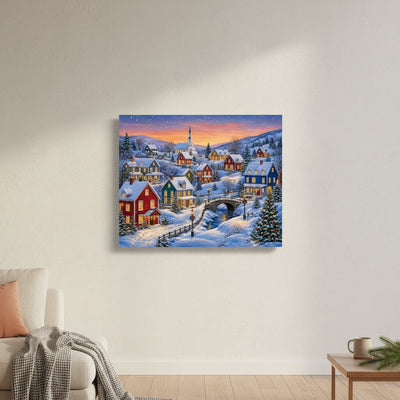 A square canvas print of a snowy village at dusk, displayed on a light beige wall in a modern living room setting with a sofa, throw blanket, and side table visible.