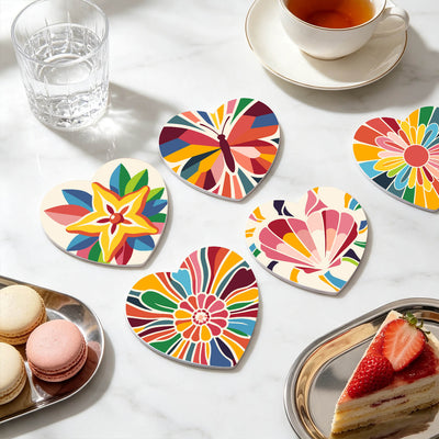 Six heart-shaped coasters with colorful, vibrant abstract and floral patterns arranged on a white marble tabletop alongside a glass of water, a cup of tea, macarons, and a strawberry cake slice. Colorful Kaleidoscope Coasters - 6 Pack Paint by Numbers Lifestyle