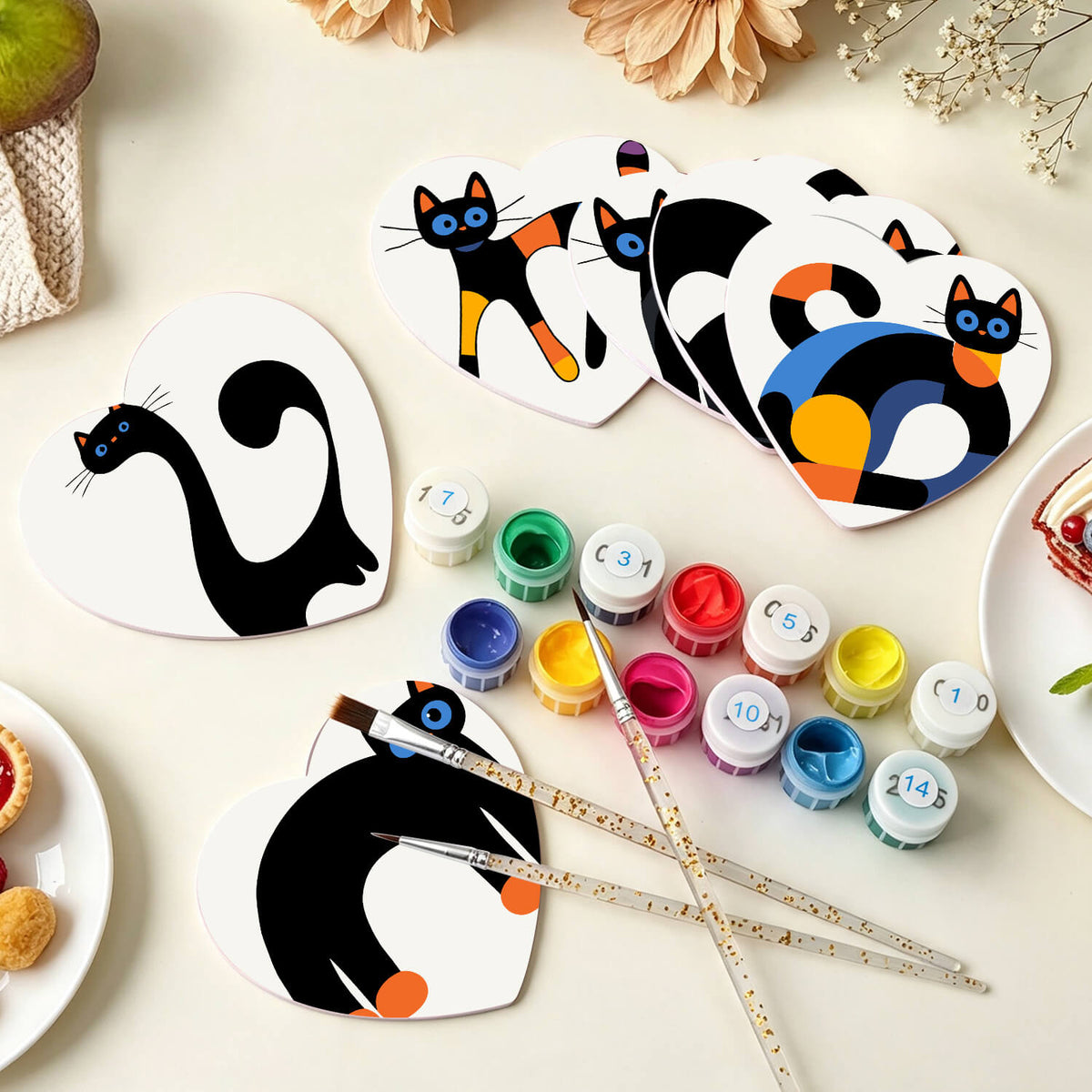 A flat-lay of heart-shaped wooden paint-by-numbers canvases featuring black and orange cat designs, shown with paint pots, paintbrushes, and plates on a light beige surface. Curious Cats Coasters - 6 Pack Paint by Numbers Package