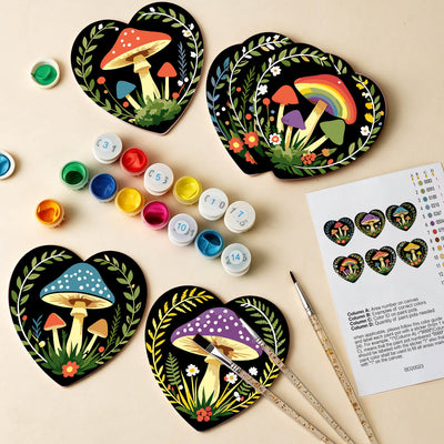 Four heart-shaped wooden coasters with hand-painted colorful mushroom designs are arranged on a light beige background with paint pots, paintbrushes, and a color chart sheet. Enchanted Mushrooms Coasters - 6 Pack Paint by Numbers Package