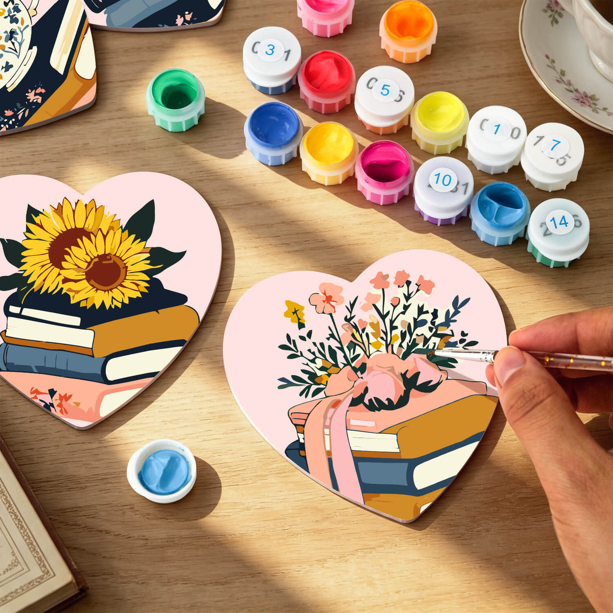 Heart-shaped paint-by-numbers art pieces on a wooden table, with one partially painted showing a sunflower on books and another being painted by a hand with a paintbrush. Color paint pots and a cup are arranged around it. Floral Blossoms Coasters - 6 Pack Paint by Numbers Package