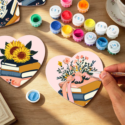Heart-shaped paint-by-numbers art pieces on a wooden table, with one partially painted showing a sunflower on books and another being painted by a hand with a paintbrush. Color paint pots and a cup are arranged around it. Floral Blossoms Coasters - 6 Pack Paint by Numbers Package
