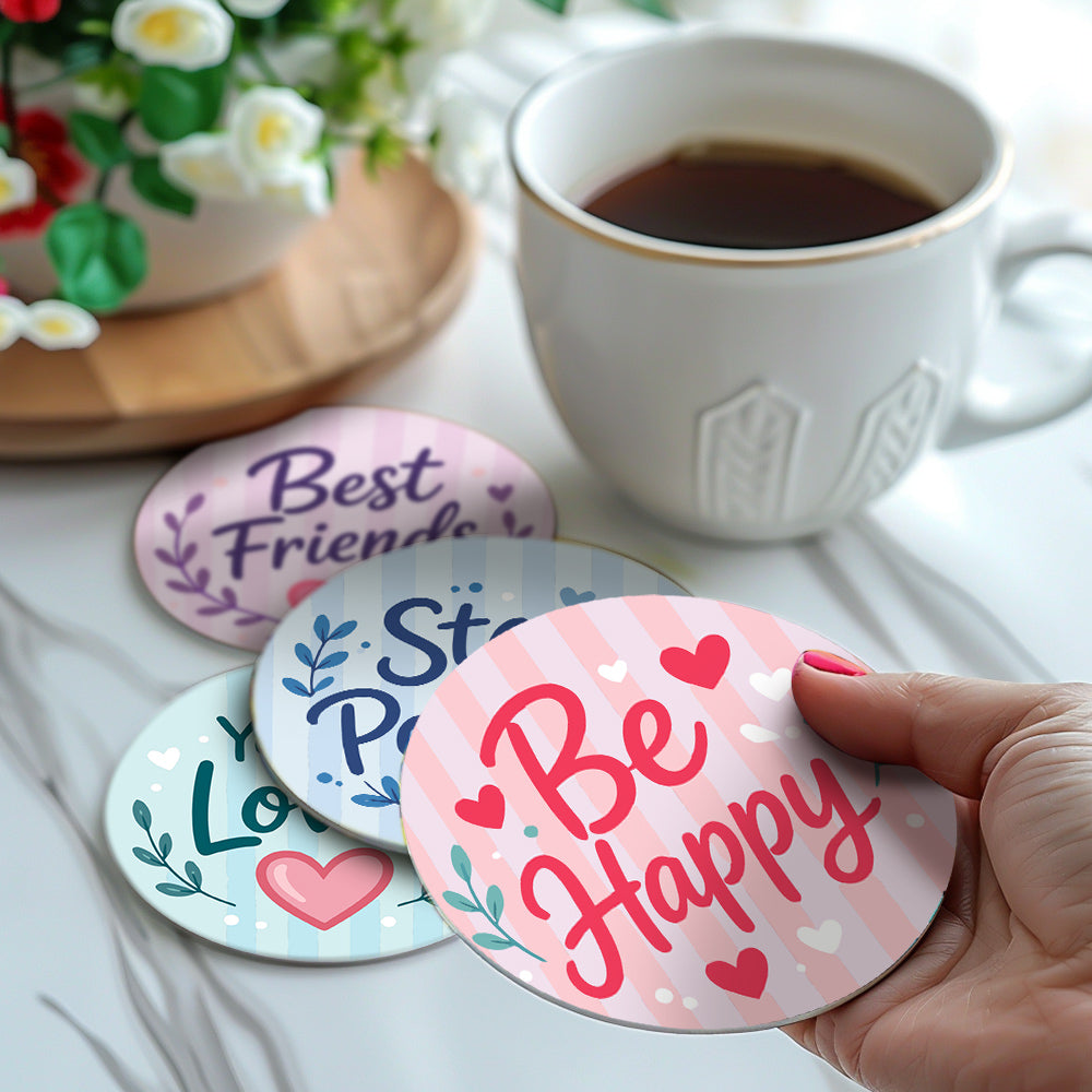 A hand holds a round coaster printed with 'Be Happy' in pink script over a pink striped background, with hearts and small floral accents. Other coasters are visible in the background, along with a white mug of coffee and a potted plant on a light marble surface. Inspiring Words Coasters - 6 Pack Paint by Numbers Lifestyle