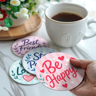 A hand holds a round coaster printed with 'Be Happy' in pink script over a pink striped background, with hearts and small floral accents. Other coasters are visible in the background, along with a white mug of coffee and a potted plant on a light marble surface. Inspiring Words Coasters - 6 Pack Paint by Numbers Lifestyle