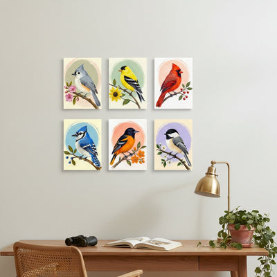Six framed bird art prints arranged in two rows on a light gray wall above a wooden desk with a lamp, book, and plant.
