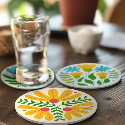 Three round coasters with colorful floral designs are arranged on a wooden table, with one coaster supporting a clear glass of water. The background is softly blurred, showing a potted plant and a small bowl. Modern Wildflowers Coasters - 6 Pack Paint by Numbers Lifestyle